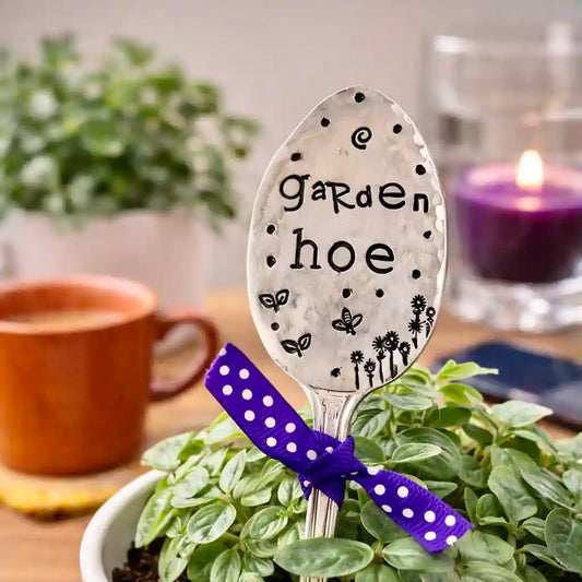 Decorative spoon with 'garden hoe' text on a plant, with a cup and candle in the background.