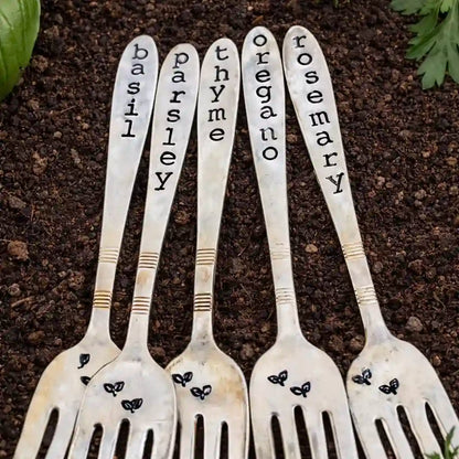 Set of five silver forks with herb names engraved on them, placed on a dark surface.
