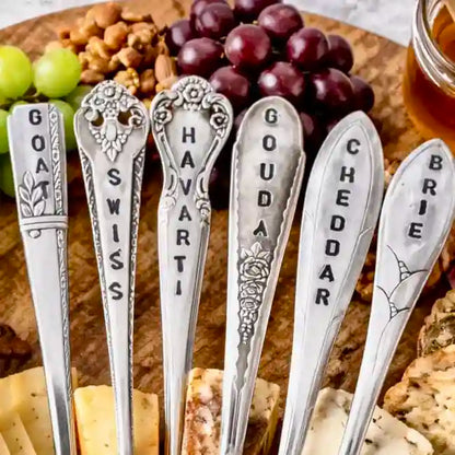 Set of silver cheese spoons with engraved names on a wooden board with grapes and crackers.