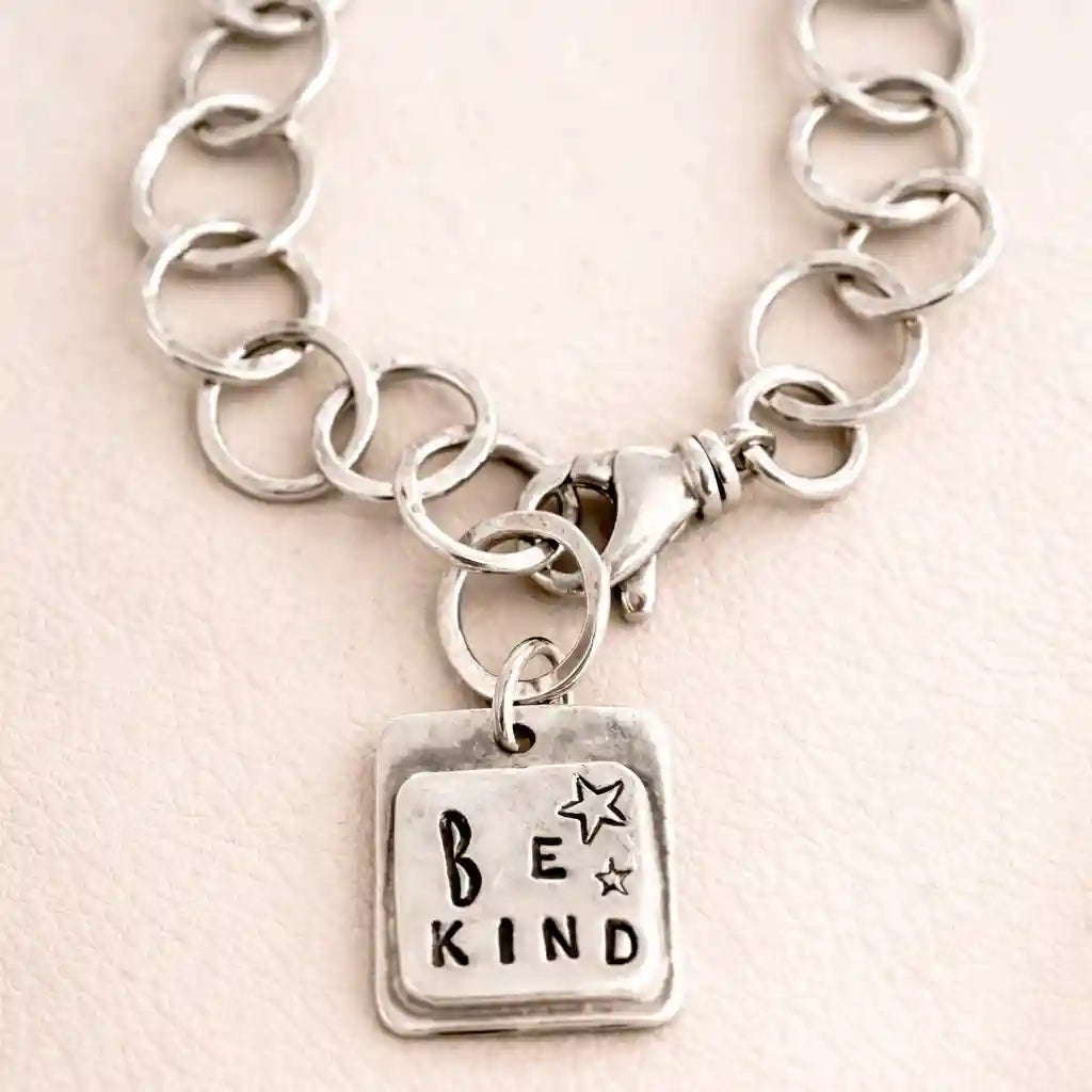 Silver chain bracelet with 'Be Kind' charm on a light pink background