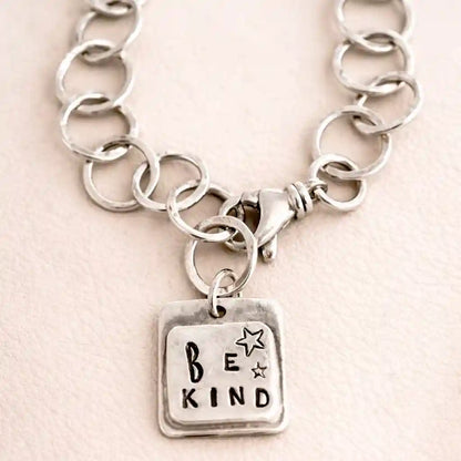 Silver chain bracelet with 'Be Kind' charm on a light pink background